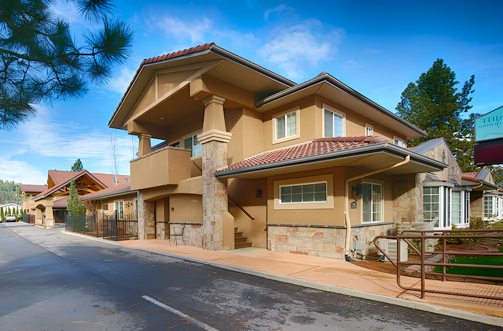 Exterior view of a senior living facility building with beige walls, stone accents, and a red-tiled roof under a blue sky with some clouds. There is a sidewalk and a ramp with a handrail leading to the entrance, and trees are visible in the background.