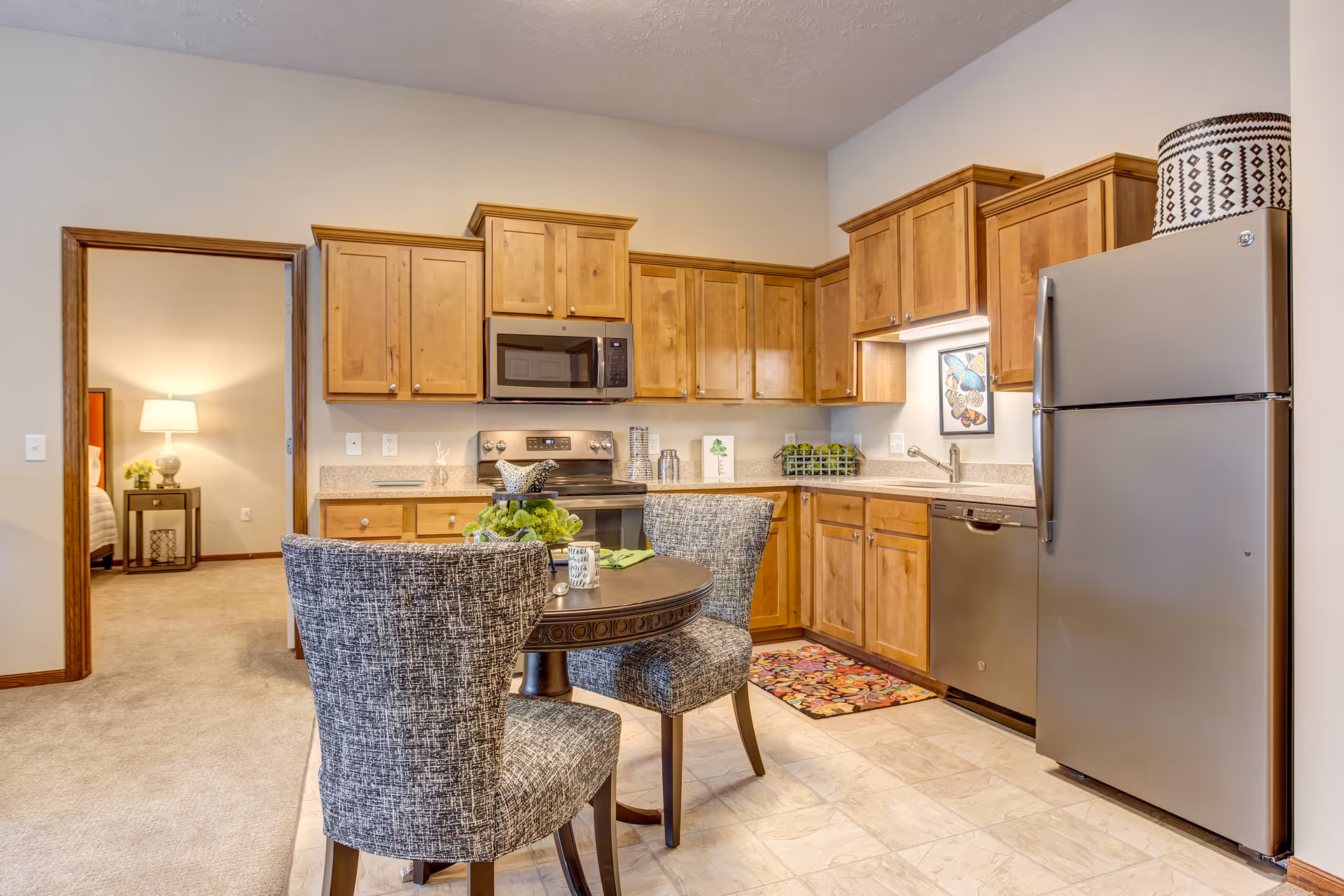 A kitchen area with wooden cabinets, a stainless steel refrigerator, stove, microwave, and dishwasher. There is a small round dining table with two upholstered chairs in the foreground. A doorway leads to a bedroom with a bedside table and lamp visible.