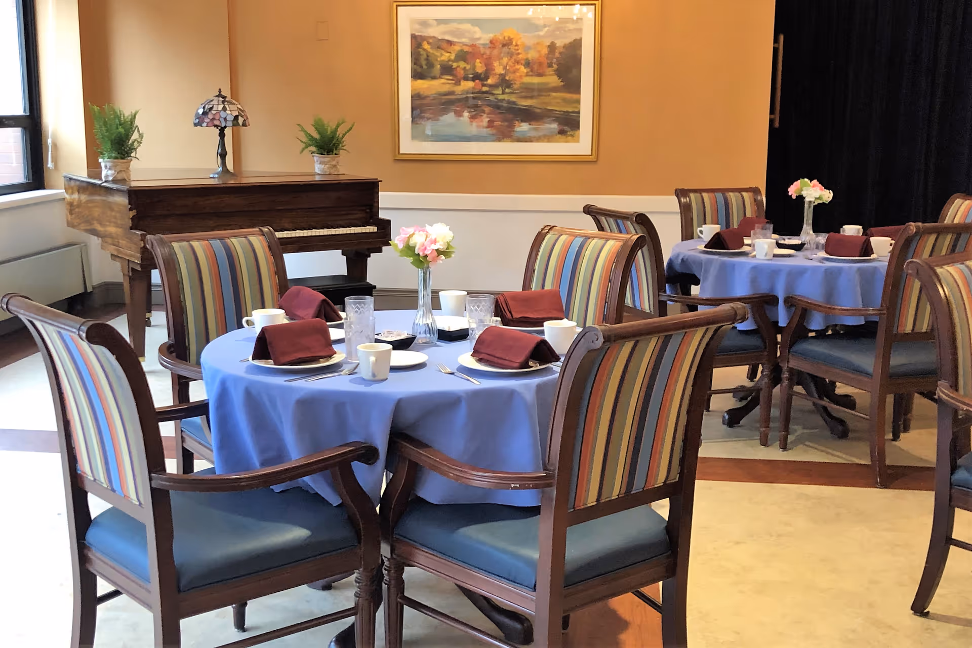 A dining room with round tables covered in blue tablecloths, set with plates, cups, glasses, and maroon napkins. Each table has a small vase with flowers. There are wooden chairs with striped upholstery around the tables. In the background, there is a wooden piano with two potted plants and a decorative lamp on top. A framed painting of a landscape with trees and a pond hangs on the wall.