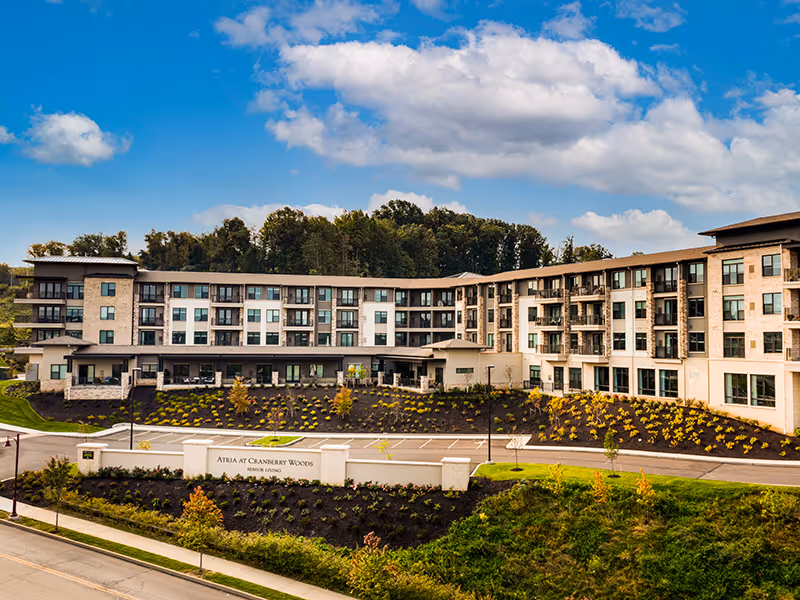 Exterior view of a large senior living facility building named Atria at Cranberry Woods, surrounded by landscaped grounds with trees and shrubs under a partly cloudy blue sky.
