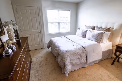 A cozy bedroom with a neatly made bed featuring a tufted headboard and floral bedding. There is a wooden dresser with decorative items on the left side and a small wooden nightstand on the right. A window with white blinds lets in natural light, and a closed door is visible next to the window.