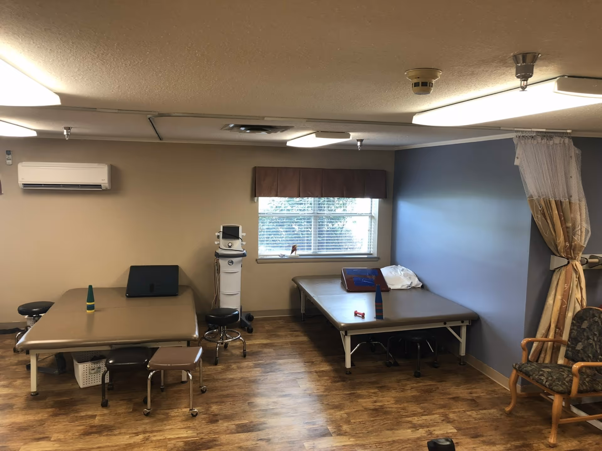 Rehabilitation therapy room with two treatment tables, stools, a medical device, window and seating.