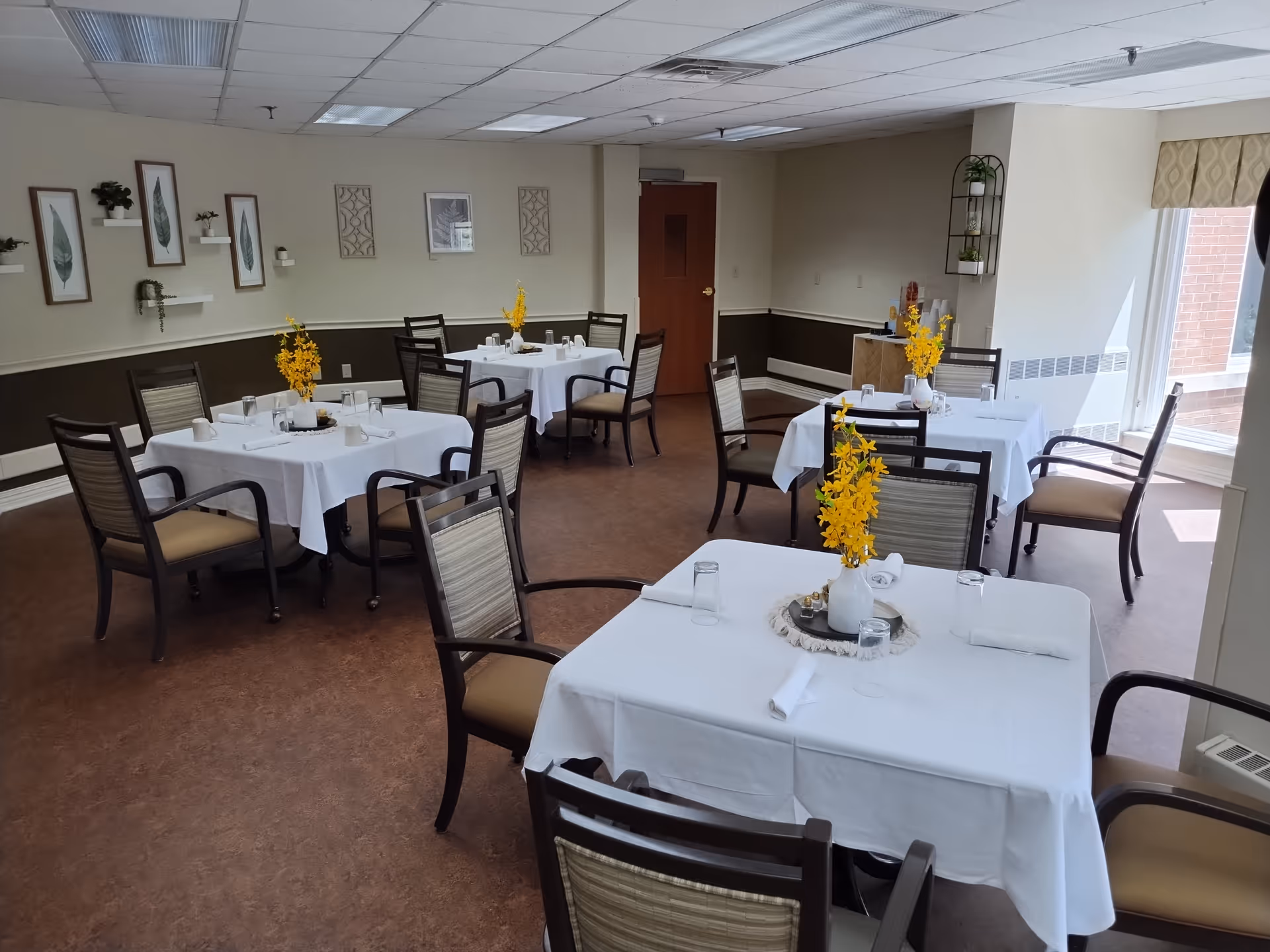 A dining room with several square tables covered with white tablecloths, each set with glasses, napkins, and a vase with yellow flowers. The room has brown flooring, beige walls with framed artwork and small shelves, and a window letting in natural light.