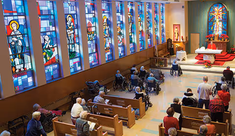 Interior view of a chapel with stained glass windows along one wall. Several elderly people, some in wheelchairs, are seated in wooden pews facing an altar decorated with flowers and religious symbols.