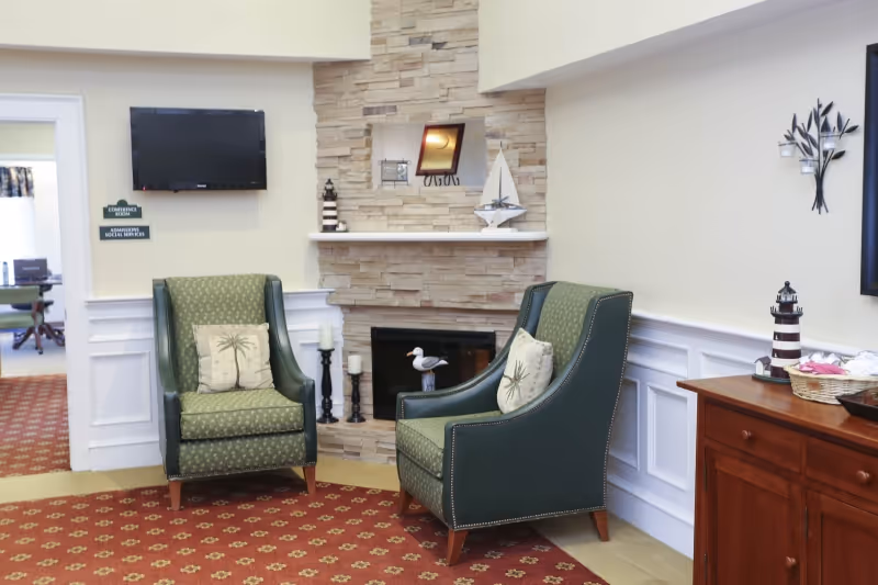 A cozy sitting area with two green upholstered armchairs featuring palm tree patterned cushions, positioned around a stone fireplace with decorative items including a model sailboat and a lighthouse figurine. A flat-screen TV is mounted on the wall above one chair, and a wooden cabinet with a basket and another lighthouse decoration is on the right. The room has white wainscoting and a red patterned carpet.