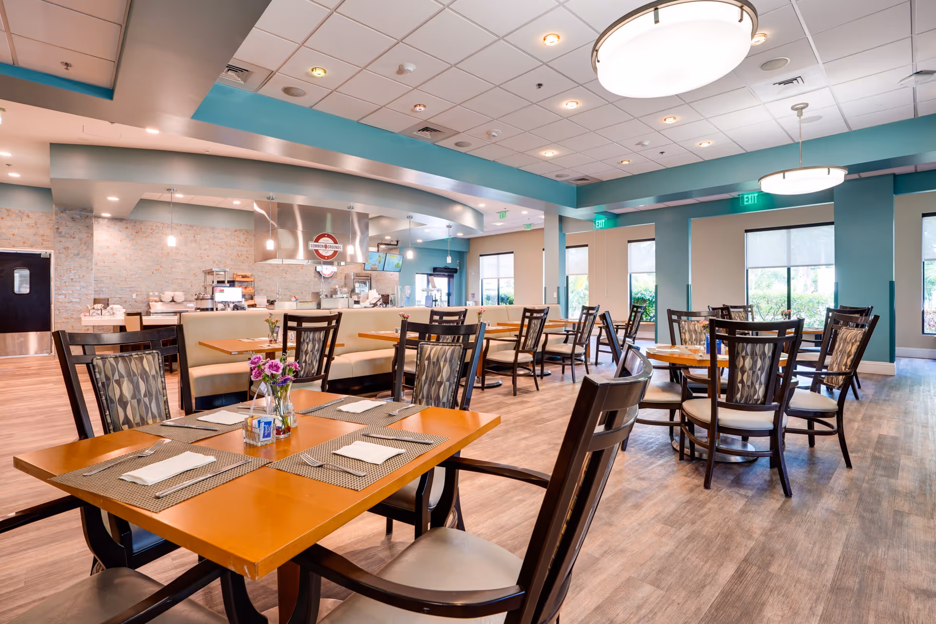 Spacious, well-lit dining room with wooden tables and chairs, place settings, and a serving counter in the background.