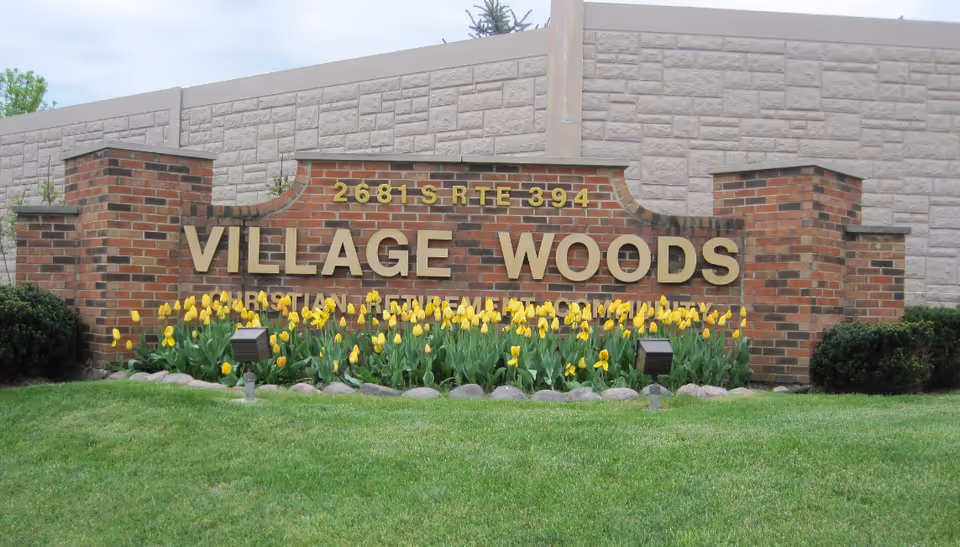 A brick sign for Village Woods, a Christian retirement community, surrounded by yellow tulips and green grass with a concrete wall in the background.