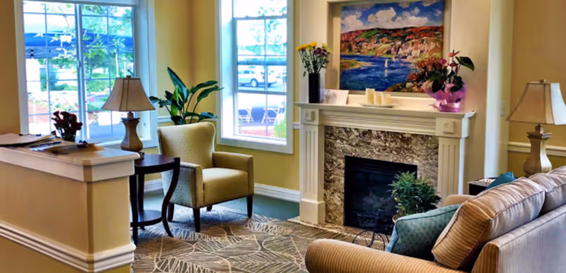 Cozy living room with armchairs and a sofa arranged around a decorative fireplace beneath a colorful painting, with large windows letting in daylight.
