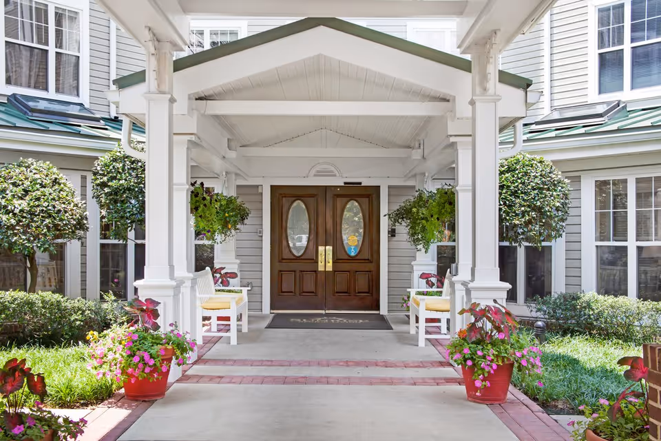 Entrance of a senior living facility with a covered porch, two wooden double doors with oval glass windows, white benches with yellow cushions on either side, hanging green plants, potted flowers, and neatly trimmed bushes and trees.