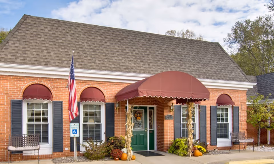 Brick single-story building entrance with maroon awnings, a green door, American flag and seasonal pumpkins at the doorway.