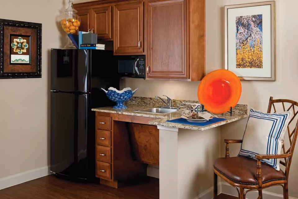 A small kitchen area with wooden cabinets, a black refrigerator, a microwave, and a granite countertop with a sink. On the counter, there is a decorative blue bowl, a large orange decorative plate, and a cup with a saucer on a blue placemat. A wooden chair with a white and blue cushion is placed next to the counter. The walls have two framed artworks.