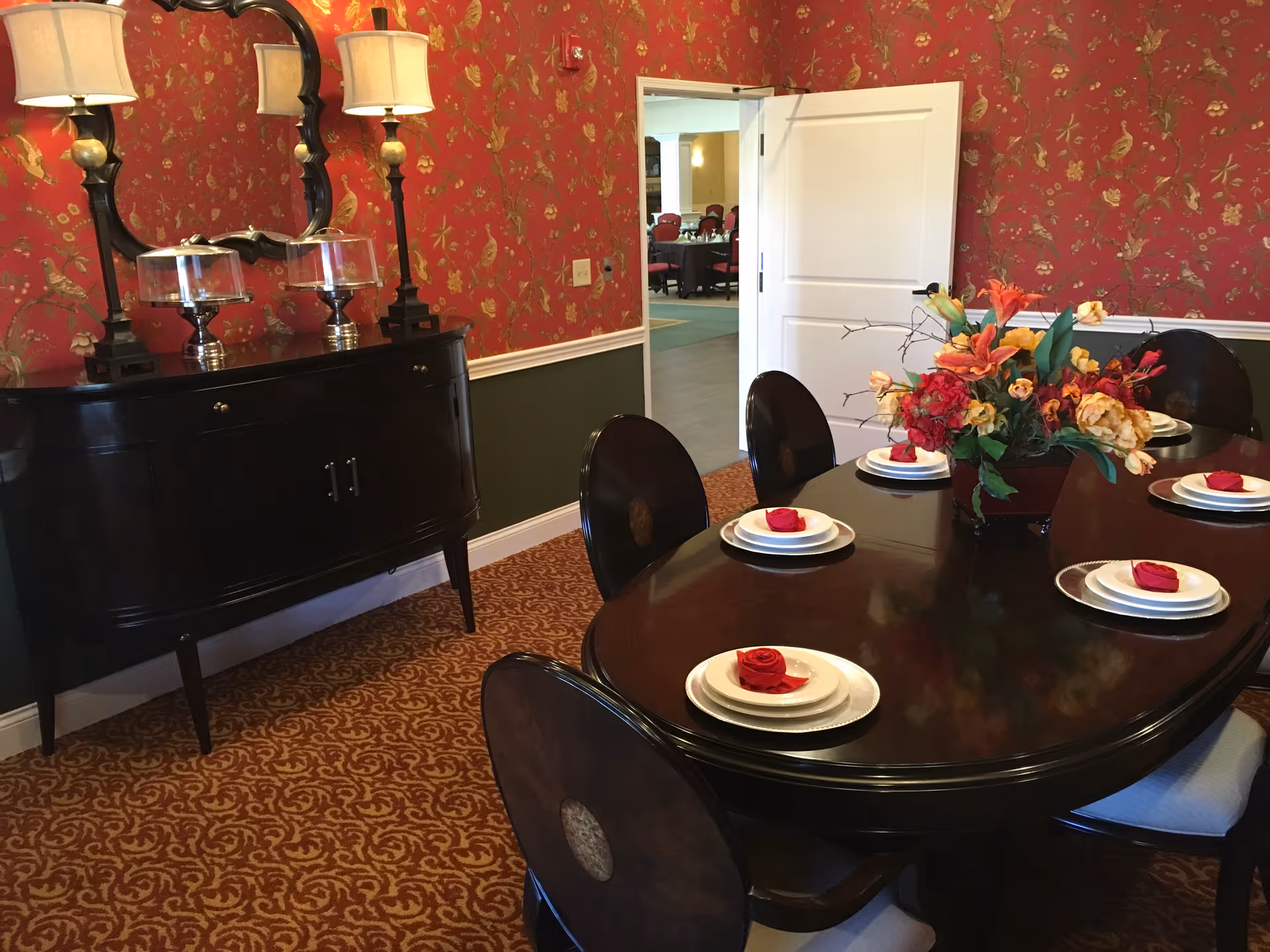 A formal dining room with a dark wooden oval table set with white plates and red napkins shaped like roses. A floral centerpiece decorates the middle of the table. The room has red wallpaper with a gold bird and floral pattern, a patterned carpet, and a black sideboard with two lamps and a mirror above it. An open door leads to another room with tables and chairs.