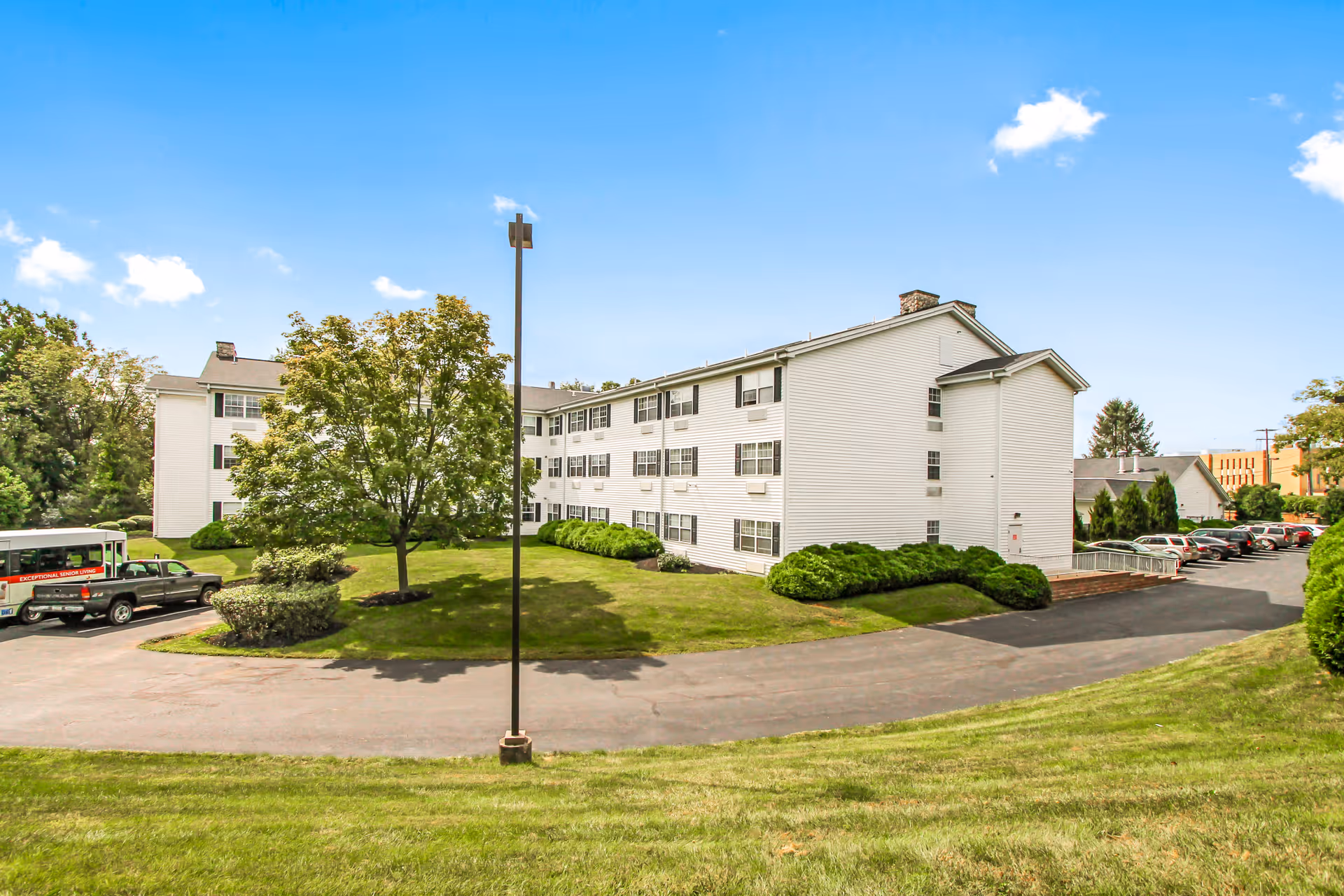 Exterior view of a three-story white residential building with multiple windows, surrounded by green grass, bushes, and trees under a clear blue sky. There is a paved driveway with parked cars and a bus nearby.
