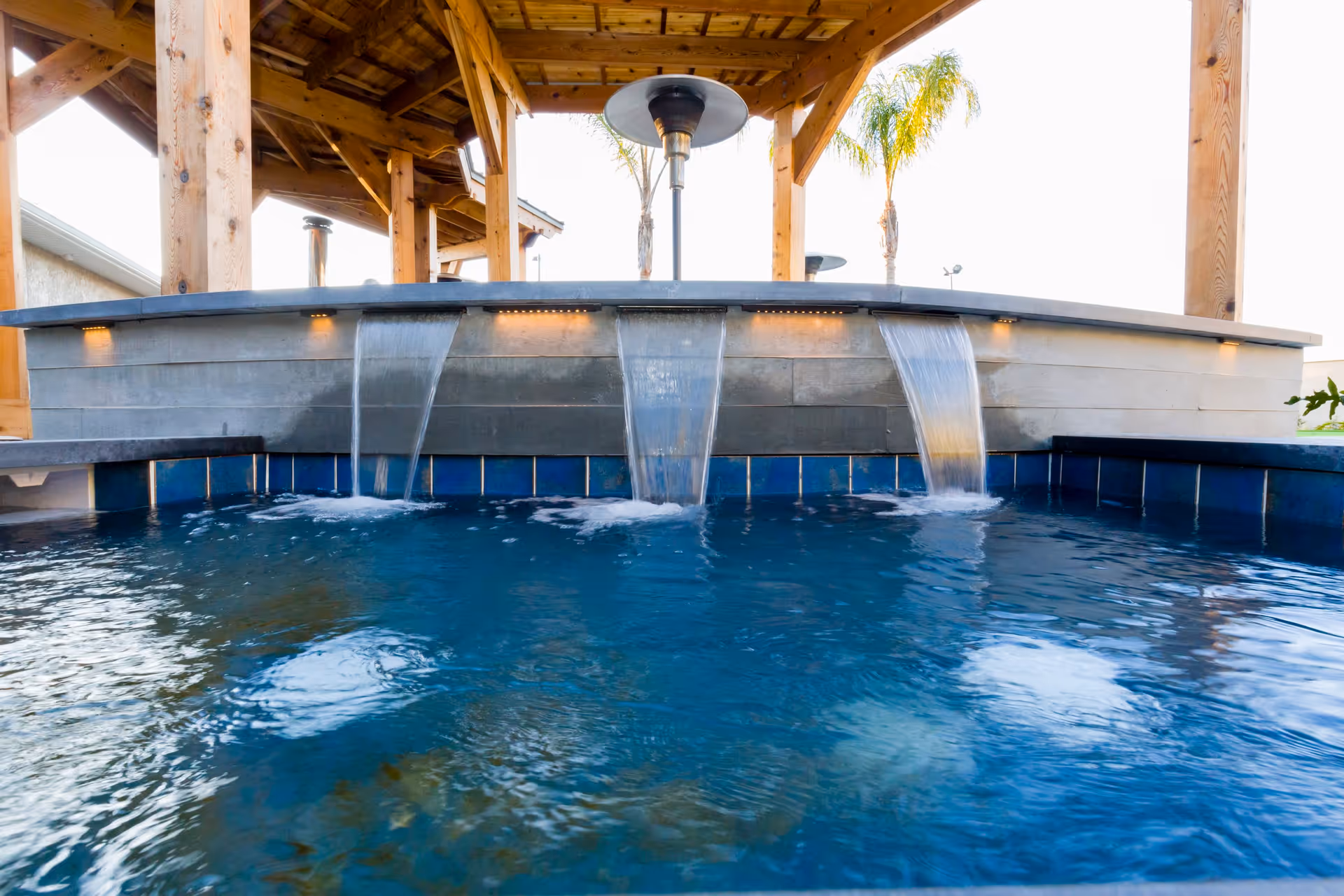 A close-up view of a water feature with three streams of water flowing into a blue-tiled pool under a wooden pergola with palm trees visible in the background.