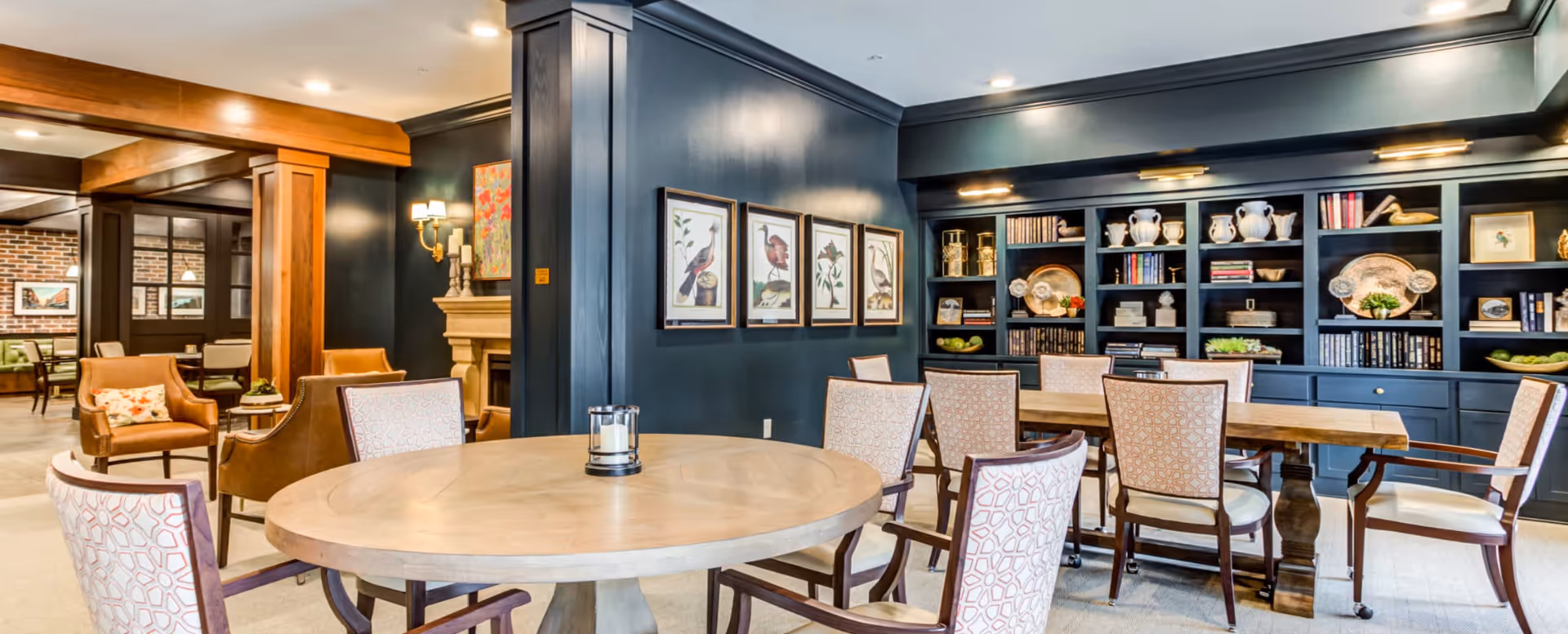 A cozy and elegant common area in a senior living facility featuring a round wooden table with six upholstered chairs in the foreground. Behind it, there is a long wooden table with additional chairs. The walls are painted dark blue and decorated with framed bird artwork and built-in shelves filled with books, decorative plates, and vases. The space is warmly lit with ceiling lights and wall sconces, and there is a fireplace partially visible in the background.