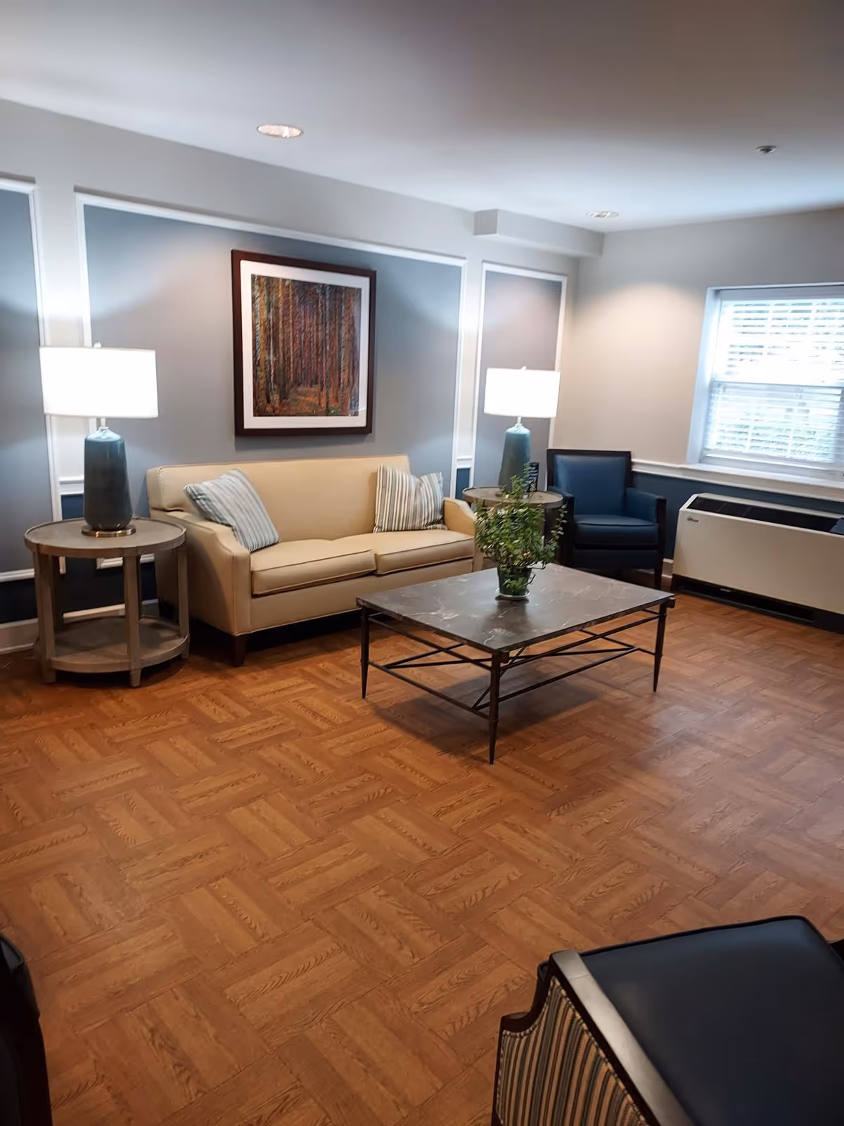 A cozy living room with a beige sofa adorned with striped pillows, two side tables each with a lamp, a dark blue armchair, a coffee table with a plant, and a framed picture of a forest on the wall. The room has wood-patterned flooring and a window with blinds.