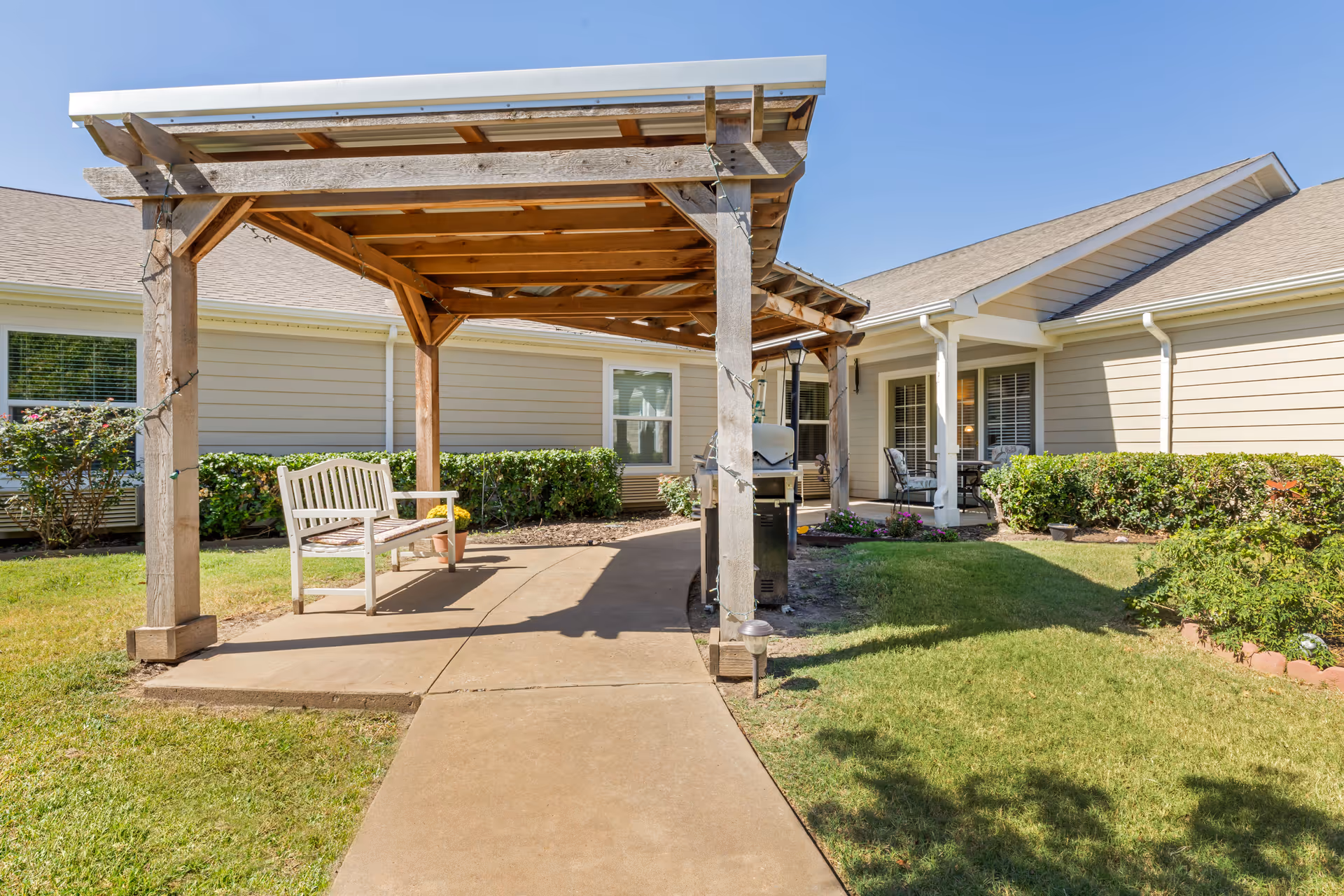 Outdoor patio area at Brookdale Owasso featuring a wooden pergola with string lights, a white bench, a barbecue grill, and a concrete pathway surrounded by green grass and shrubs. The building exterior is beige with white trim and windows.