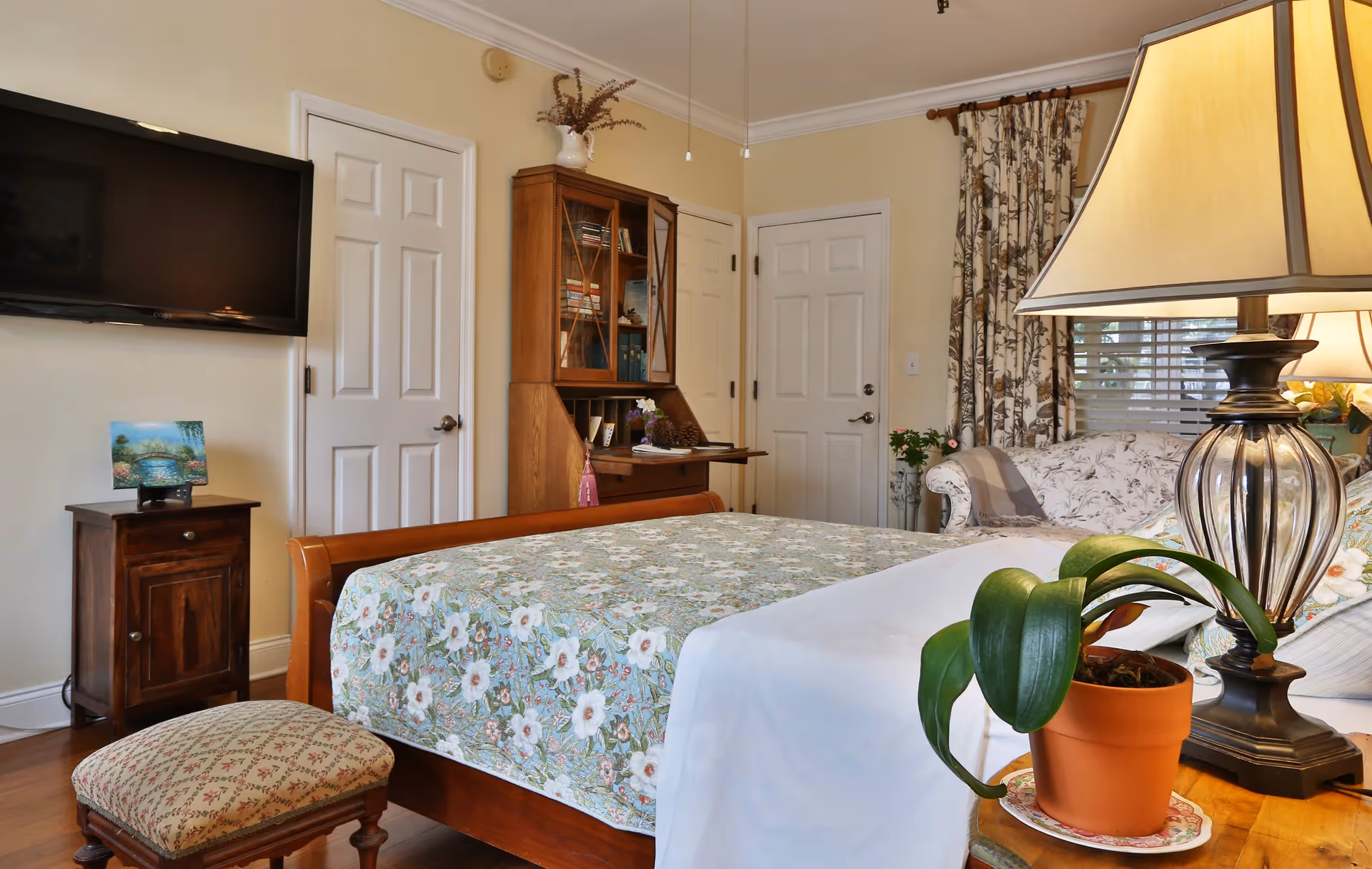 Furnished bedroom with a floral bedspread, wooden sleigh bed, bedside lamp and plant, cabinets, and a wall-mounted TV.