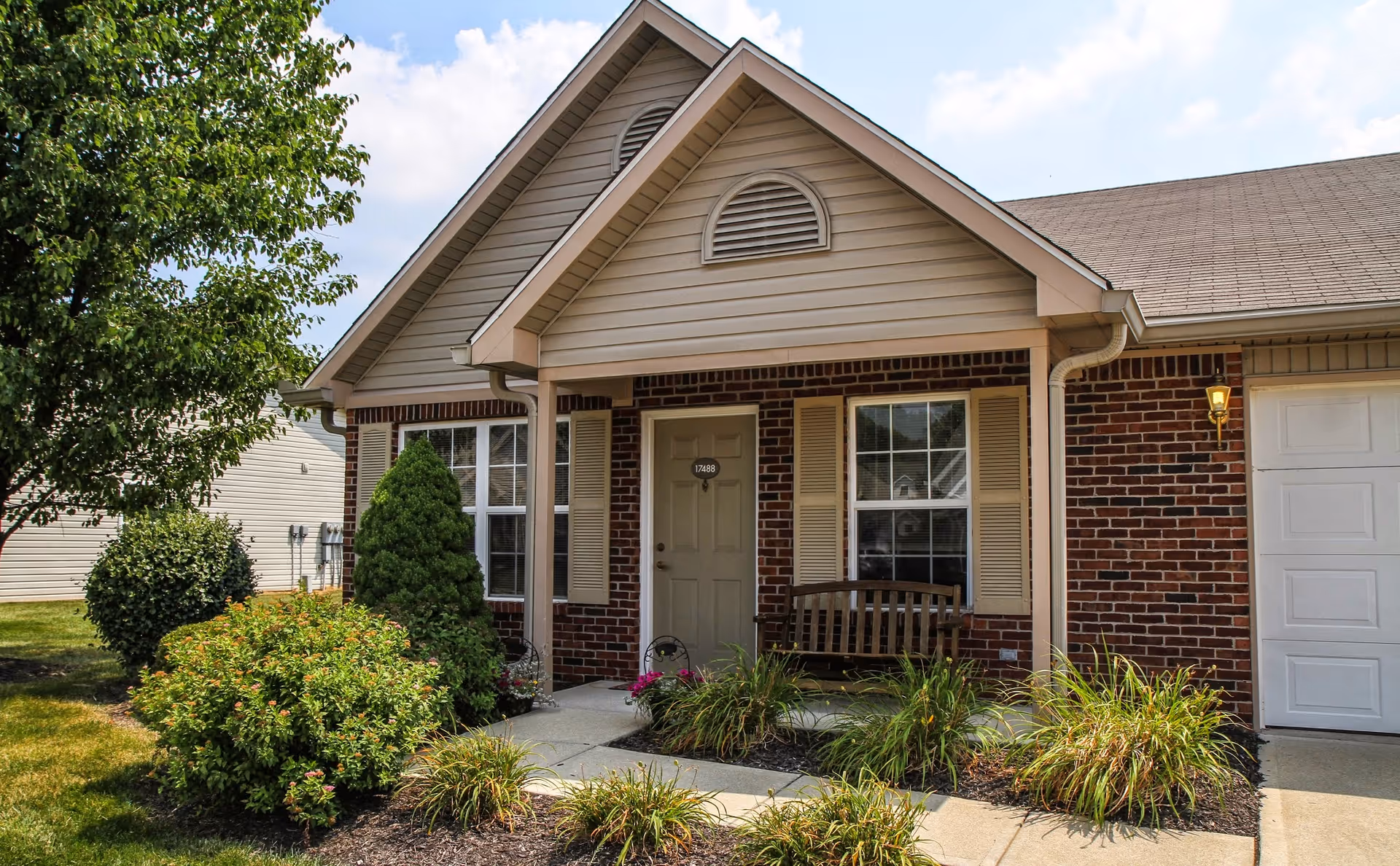 Front entrance of a single-story brick residence with a covered porch, bench, and landscaped shrubs beside a garage.