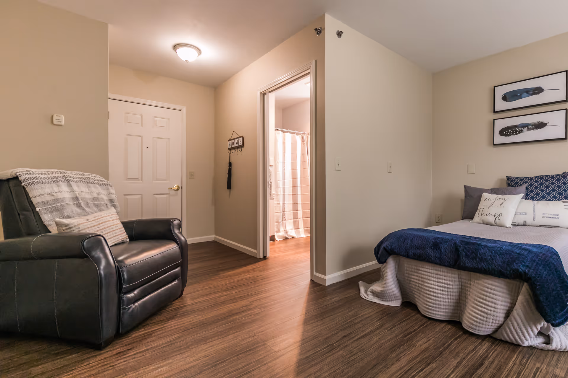 A furnished studio-style bedroom with a bed, armchair, wood flooring, and a doorway leading to a bathroom.