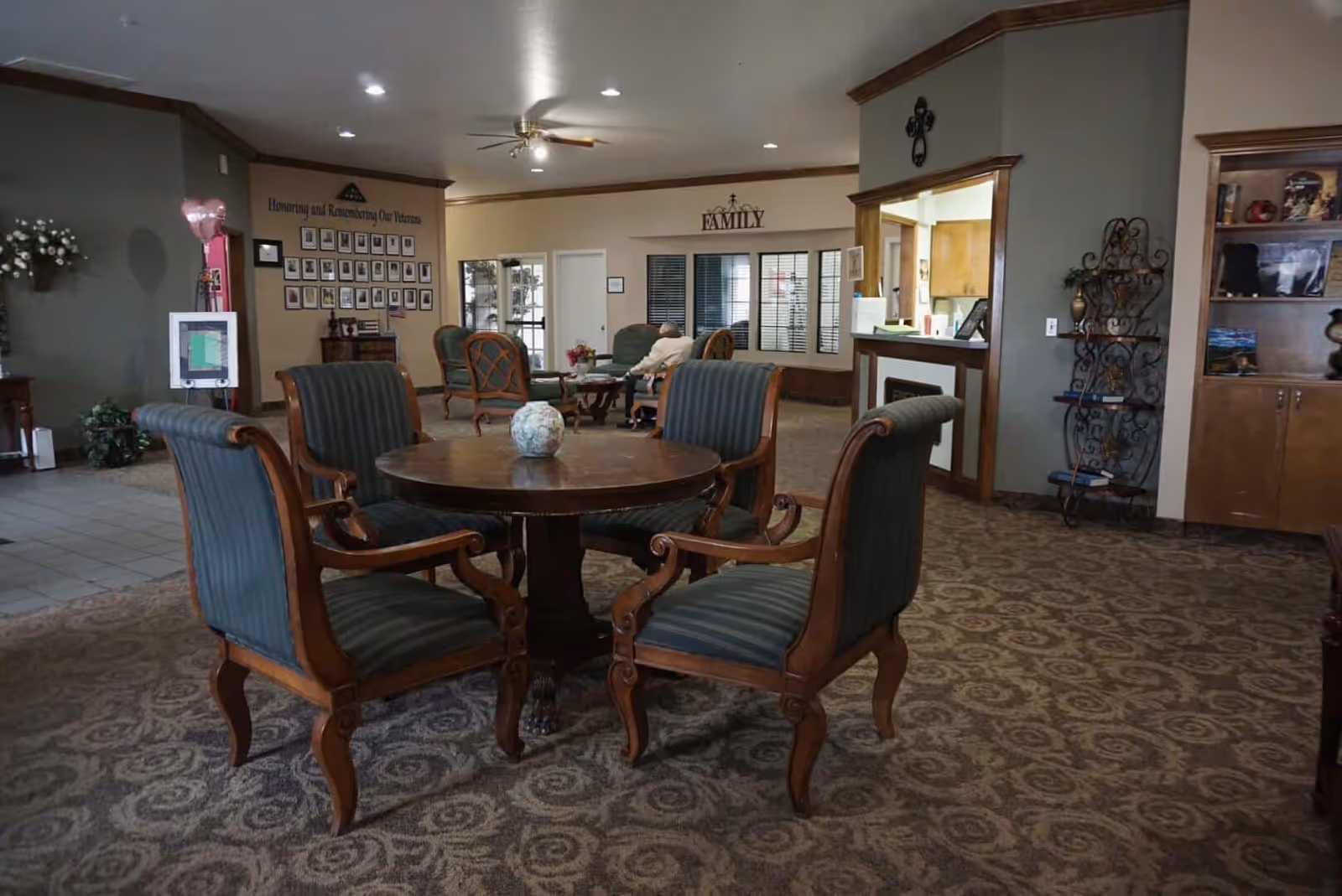 A senior living facility common area with a round wooden table surrounded by four upholstered chairs in the foreground. In the background, there are additional seating areas with armchairs, a wall display honoring veterans, and a sign that reads FAMILY above a windowed office area. The room has carpeted floors, recessed lighting, and decorative wood trim.