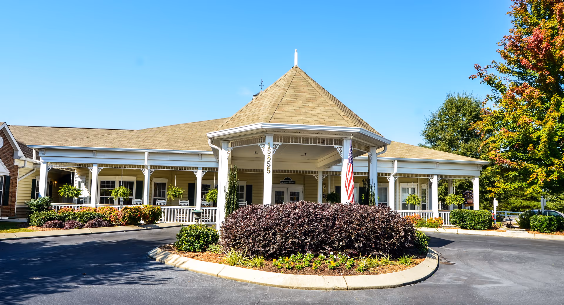 Front exterior view of a single-story building with a large covered porch supported by white columns. The building has a tan roof and beige siding, with several hanging green plants along the porch. There is a circular driveway with landscaped bushes and flowers in the center. An American flag is displayed near the entrance. Trees with green and autumn-colored leaves are visible in the background under a clear blue sky.