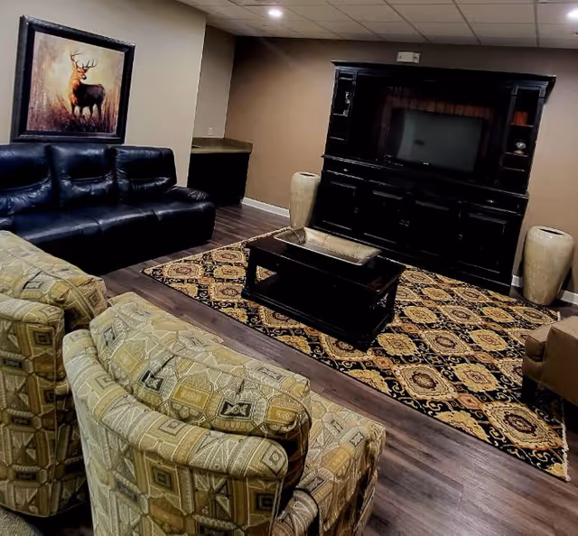 A cozy living room with a patterned area rug, a black leather couch, two patterned armchairs, a dark wood coffee table, and a large dark wood entertainment center with a flat-screen TV. There is a painting of a deer on the wall and two large decorative vases on either side of the entertainment center.