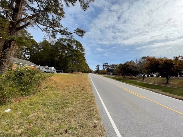 A two-lane road with a grassy shoulder on the left side and trees lining both sides. There are some parked vehicles and a building partially visible on the left side. The sky is partly cloudy.