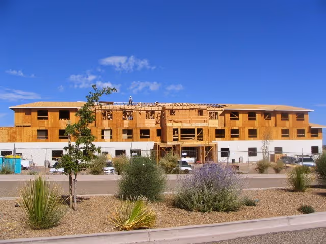A multi-story building under construction with wooden framing and partial exterior walls. The building is surrounded by a landscaped area with bushes, small trees, and desert plants under a clear blue sky.