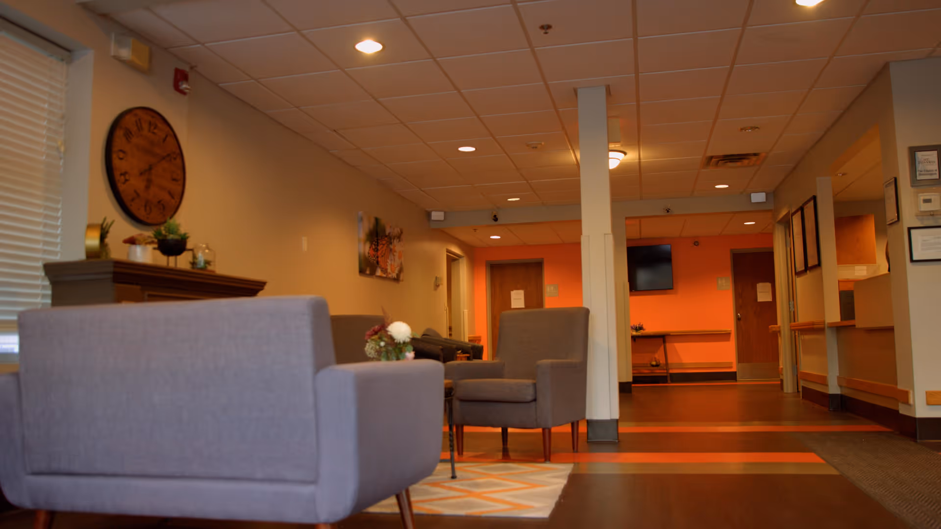 Interior view of a senior living facility lounge area with two gray armchairs, a small table with flowers, a large wall clock, and a hallway leading to doors and a wall-mounted TV.