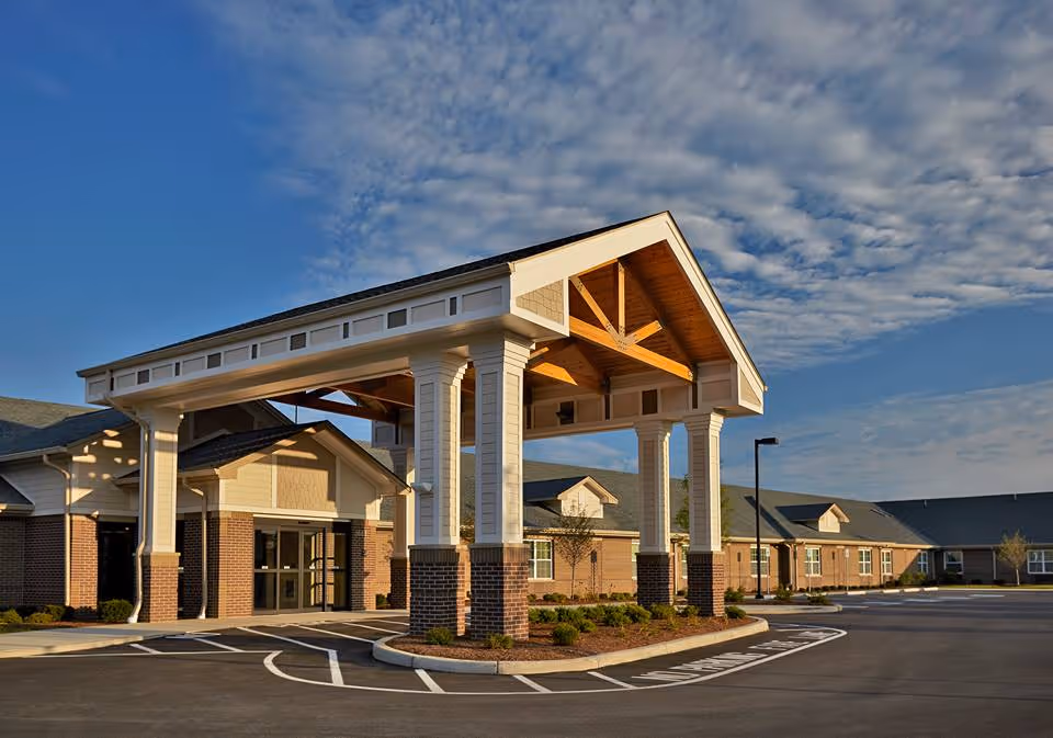 Exterior view of Harbour Manor & The Lodge building entrance with a large covered drop-off area supported by white columns with brick bases, surrounded by a parking lot and landscaping under a partly cloudy sky.