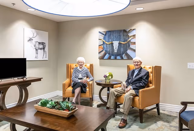 An elderly man and woman sitting in orange armchairs in a well-lit living room with beige walls. There is a wooden coffee table with a planter of succulents in front of them, a round side table with a small flower arrangement between them, and two pieces of wall art featuring a deer and a bull skull. A flat-screen TV is on a wooden stand to the left.