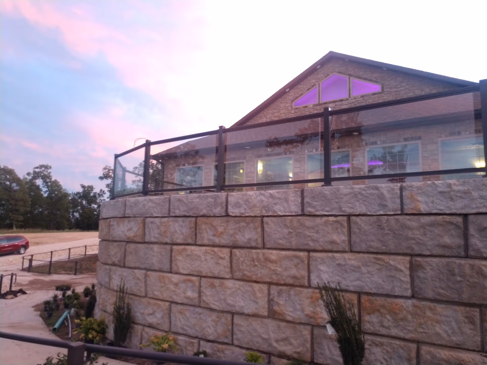Exterior view of a senior living facility building with stone walls and a glass railing on an elevated patio. The sky is at dusk with pink and blue hues, and there are trees and a parked red car in the background.