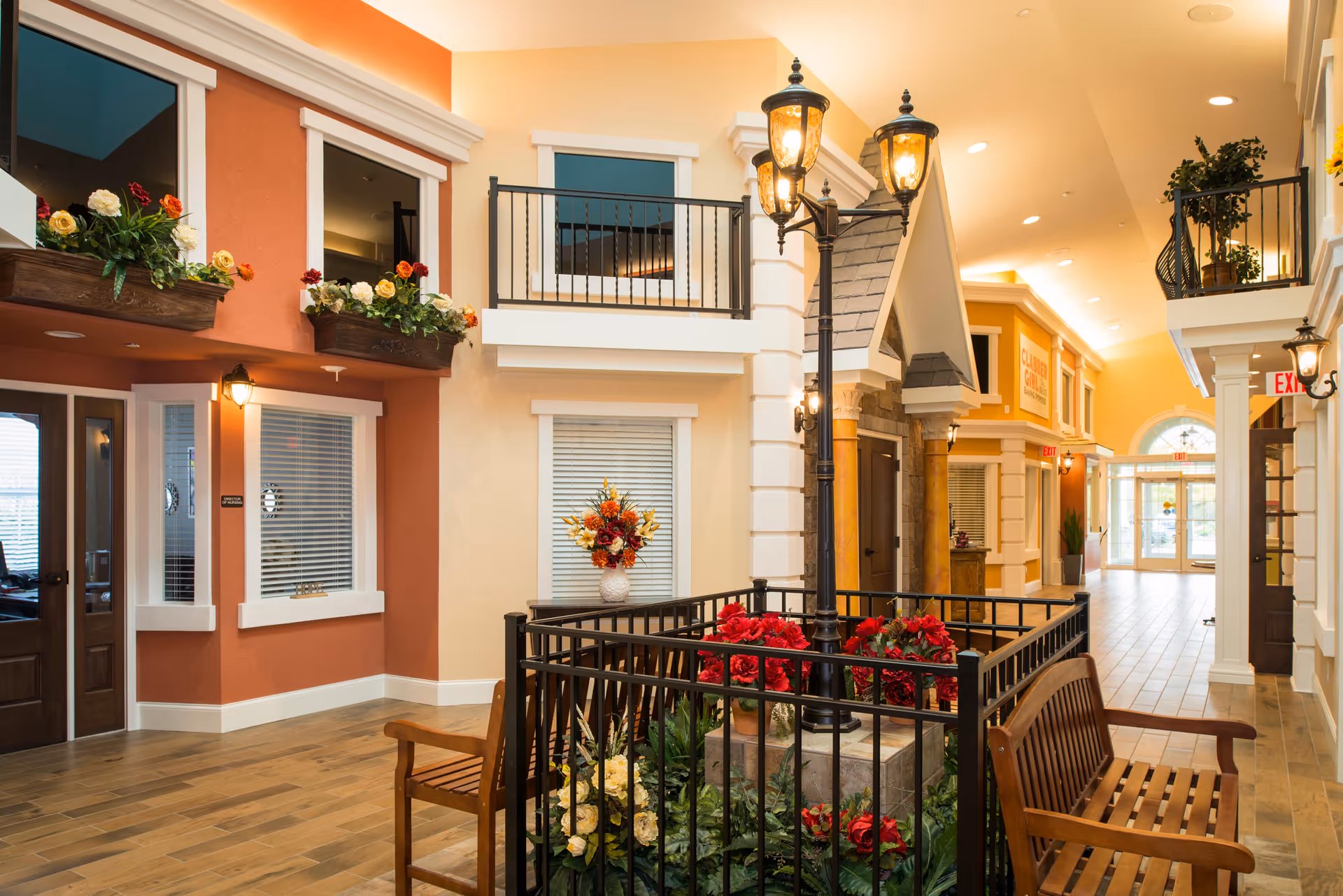 Indoor hallway designed to look like an outdoor street scene with building facades, flower boxes, a black metal fence enclosing a garden with red and yellow flowers, wooden benches, and vintage-style street lamps. The hallway has warm lighting and a tiled floor, with doors and windows along the sides and an exit door visible at the far end.