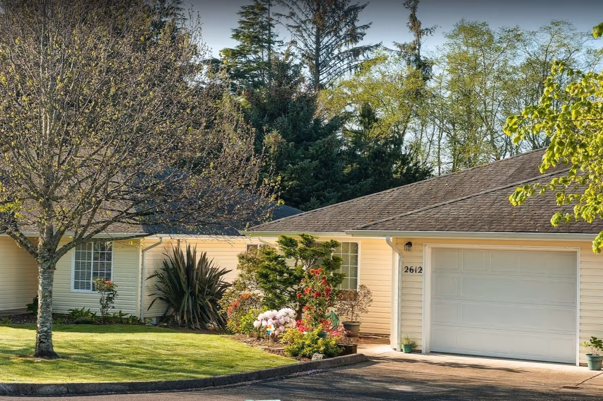 Exterior view of a single-story residential building with light yellow siding, a gray shingled roof, a white garage door, and the number 2612 on the wall. The front yard has a well-maintained lawn, a tree with budding leaves, and various shrubs and flowering plants. Tall evergreen trees are visible in the background under a clear sky.