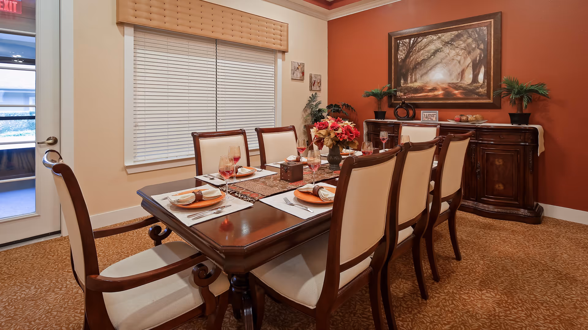 A formal dining room with a dark wooden table set for six people. The table has cream-colored placemats, orange plates, folded napkins, and wine glasses with red liquid. A floral centerpiece is in the middle of the table. The room has beige walls with one accent wall painted dark red, decorated with a large framed painting of a tree-lined path. There is a wooden sideboard against the red wall with decorative plants and items on top. A window with closed blinds and a beige valance is on one wall, and a door with a window is visible to the left.