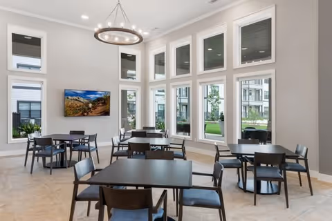 A bright dining area with multiple black square tables and matching chairs arranged neatly. The room features tall windows allowing natural light to fill the space, and a modern circular chandelier hanging from the ceiling. A flat-screen TV is mounted on the wall displaying a scenic landscape.