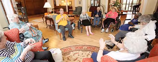 A group of elderly people sitting in a circle in a spacious, well-lit living room with wooden floors and comfortable chairs, engaging in a group activity or conversation.