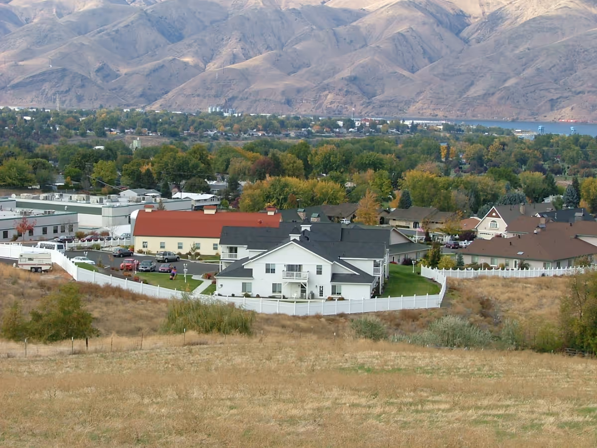 Aerial view of a senior living facility named Generations at Lewiston, showing multiple buildings with white fences, parking areas with cars, surrounded by trees and hills in the background.