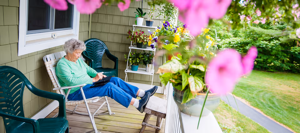 An elderly woman sitting on a porch in a white chair with her feet resting on a small wooden stool. She is wearing a green long-sleeve shirt and blue jeans, and is looking down at a tablet or book in her hands. The porch has green plastic chairs, potted plants on a white shelf, and colorful flowers in the foreground. The background shows a green lawn and trees.