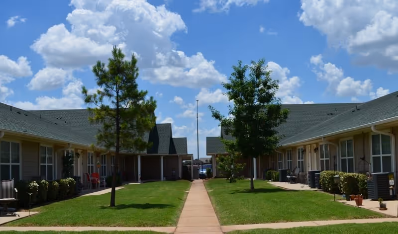 Straight paved walkway between two single-story senior living buildings with grass, small trees, and a bright blue sky with clouds.