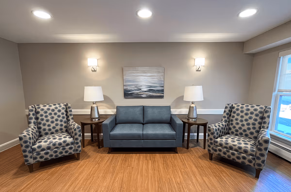 A cozy seating area in a senior living community featuring a blue loveseat sofa flanked by two patterned armchairs with blue circular designs. Each side of the sofa has a wooden side table with a silver lamp. Above the sofa is a painting depicting an abstract ocean scene. The room has wooden flooring, beige walls, two wall-mounted lights, and a window on the right side letting in natural light.
