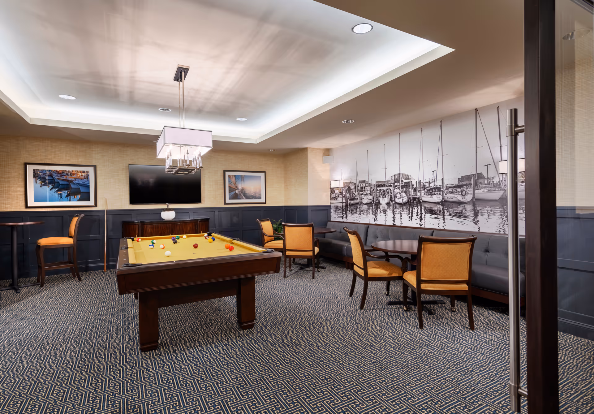 A recreational room with a pool table in the center, surrounded by seating areas including chairs and a long cushioned bench. The room has a patterned carpet, framed pictures on the walls, a flat-screen TV, and a large black and white mural of boats docked at a marina. The ceiling features recessed lighting and a modern chandelier above the pool table.
