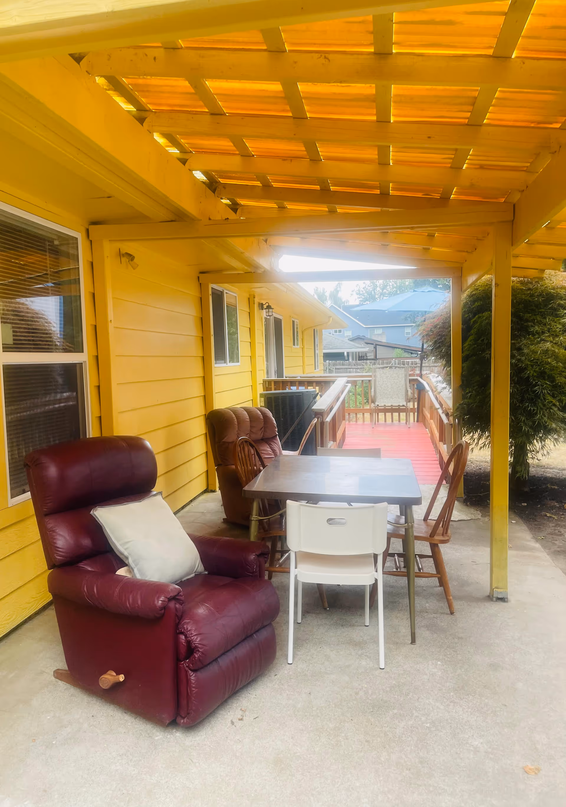 Covered outdoor patio area with a yellow wooden exterior wall, featuring a maroon recliner chair with a white pillow, a brown armchair, a table, and several chairs. The patio has a wooden pergola roof and overlooks a red wooden ramp leading to a fenced yard with neighboring houses visible in the background.