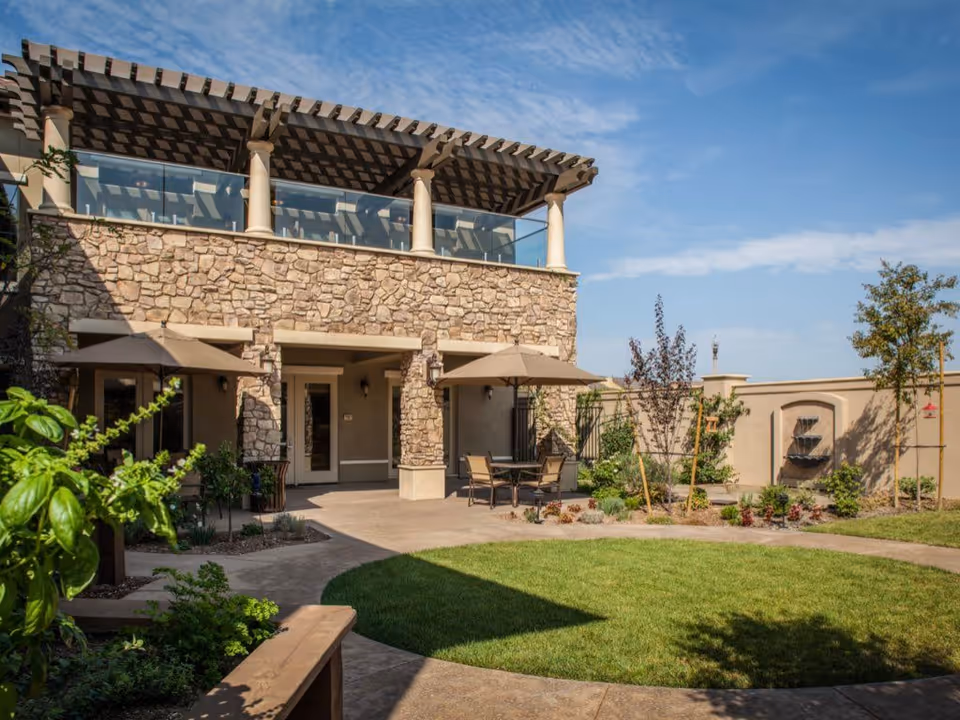 Stone-faced memory care courtyard with patio seating and umbrellas, a pergola balcony, lawn, and landscaping under a blue sky.