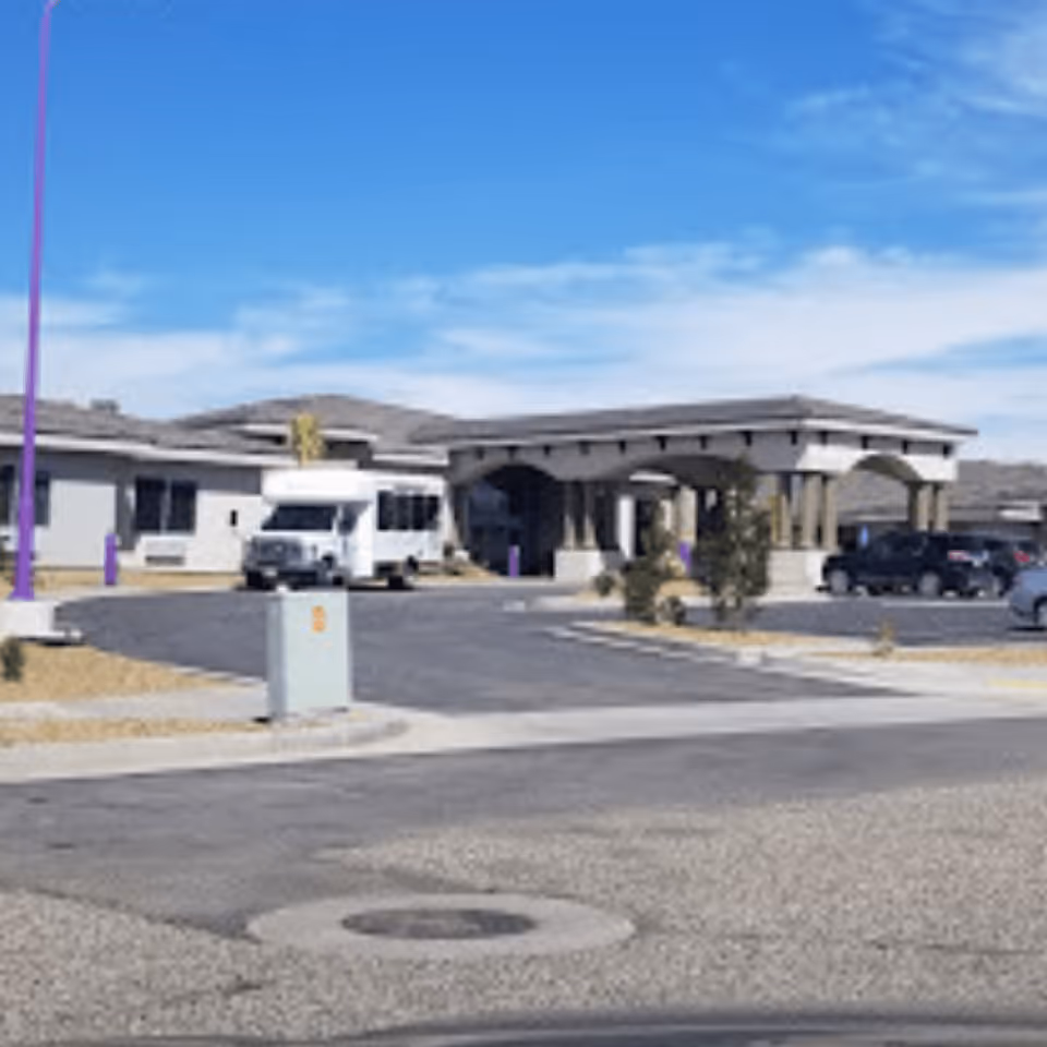 Front entrance and driveway of a senior living facility with a covered porte-cochère, a parked shuttle van and cars under a clear blue sky.