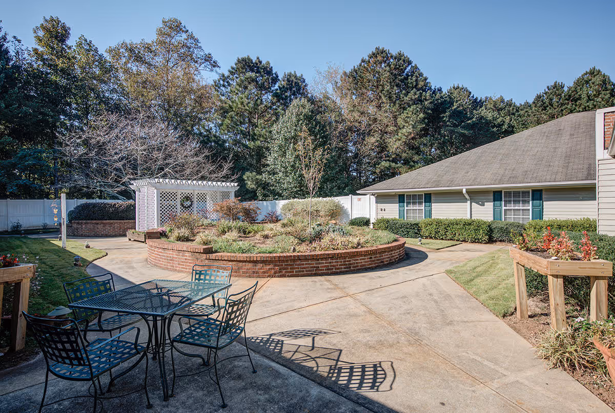 Outdoor courtyard with a metal table and chairs, a circular raised brick planter, a white lattice pergola, and a one-story building surrounded by trees.