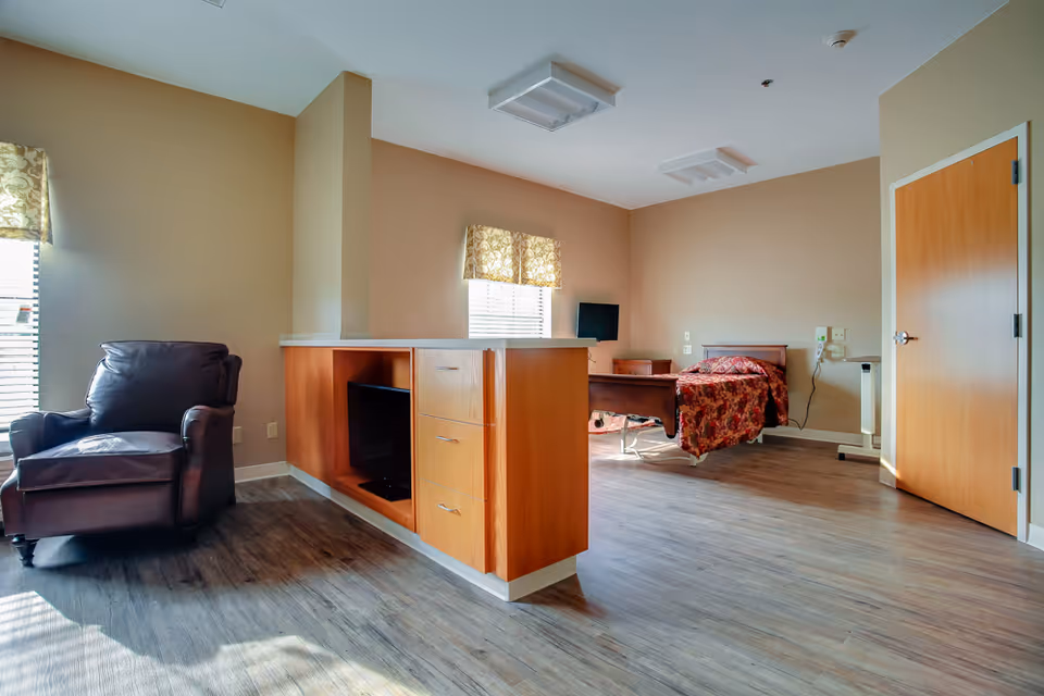 A spacious room in a senior living facility with a single bed covered in a red floral bedspread, a wooden nightstand, a small wall-mounted TV, a brown leather armchair, and a wooden cabinet with drawers and a shelf. The room has beige walls, two windows with patterned valances, and wood-look flooring.