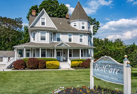 Front exterior of a Victorian-style BestCare Assisted Living house with a sign and lawn.