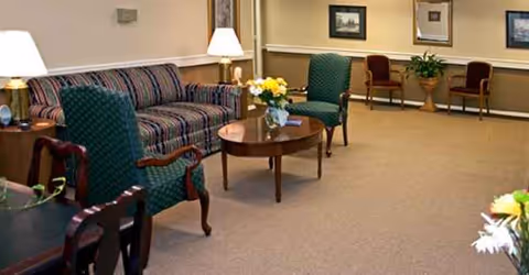 Cozy senior living lounge with striped sofas, chairs, coffee tables, lamps, and flowers.